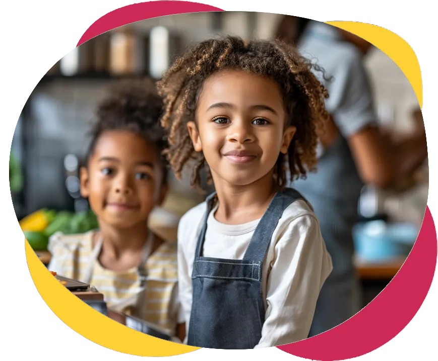 Image de deux filles souriantes assises à une table dans une cuisine, la mère des filles est derrière elles en train de préparer à manger sur la cuisinière. Image of two smiling girls sitting at a table in a kitchen, the girls' mother is behind them preparing food on the stove.