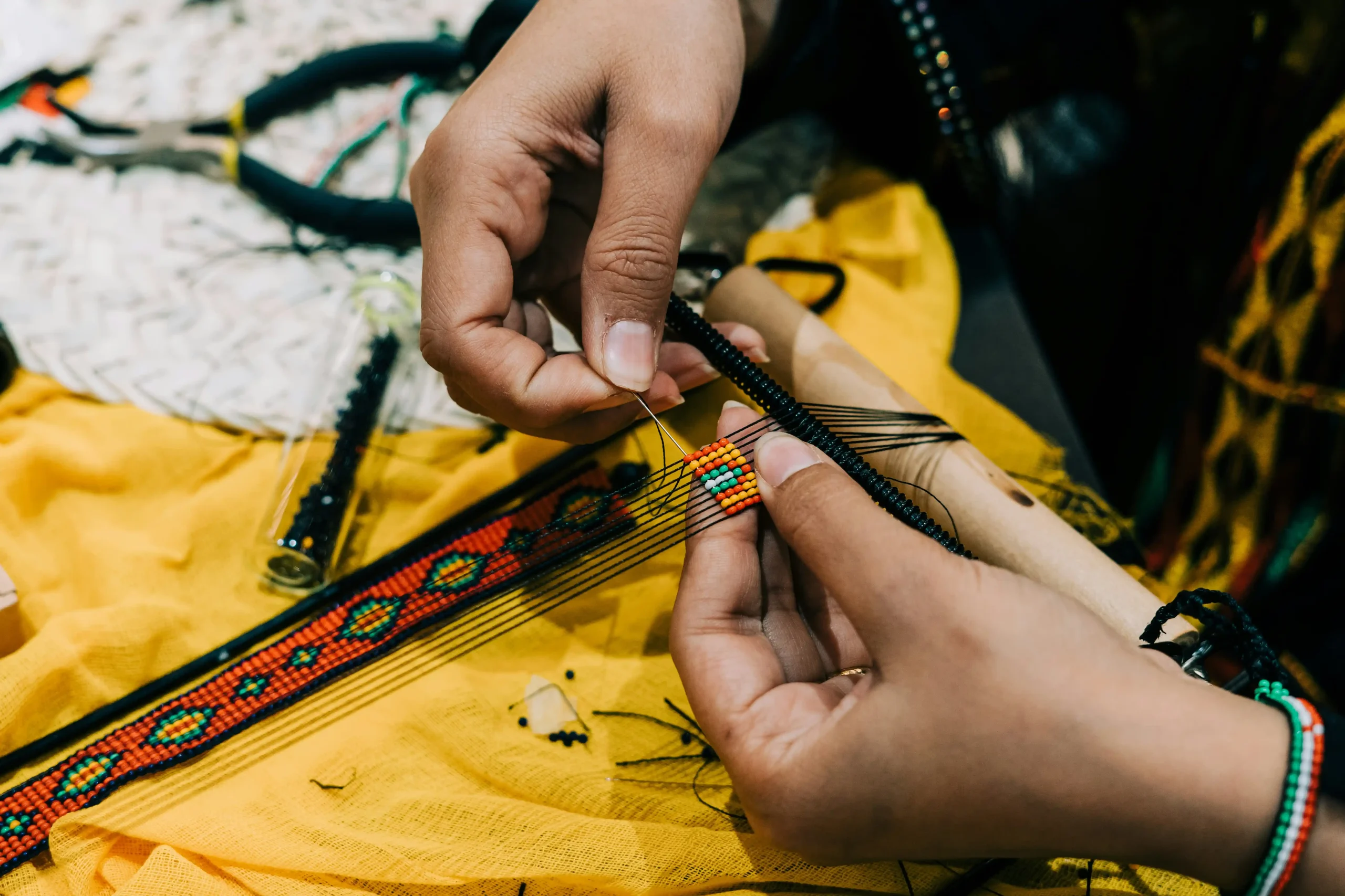 La Ruche d’art Kokomino-Mission inclusion les mains d’une personne attentivement en train de tisser un bracelet coloré avec des perles sur un métier à tisser.