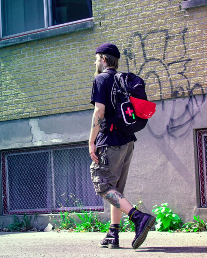Un homme avec un sac à dos et une trousse de premiers soins rouge marche le long d'un mur en briques de couleur jaune verdâtre avec des graffitis.