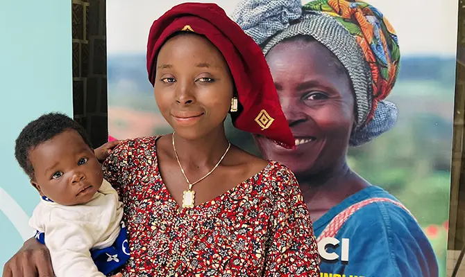 Une femme portant une tenue traditionnelle rouge tient un bébé dans ses bras, devant une affiche montrant une autre femme souriante en habits colorés.